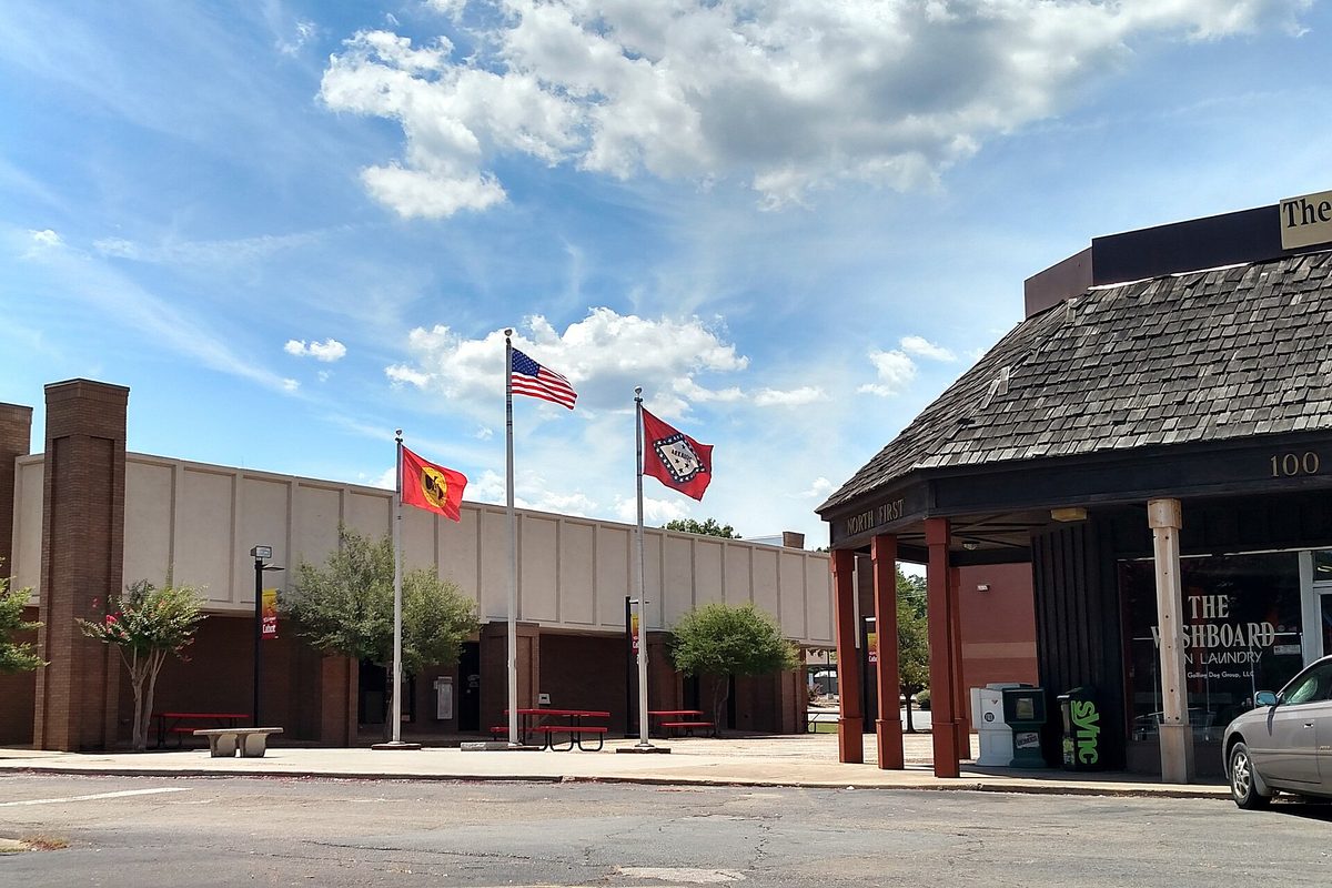US, Arkansas and red Cabot city flags on poles outside a tan and brick building downtown, next to a shingle-roofed laundromat