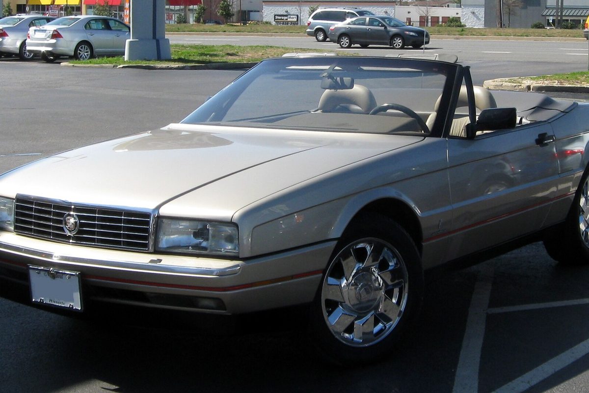 A champagne Cadillac Allante two-seat convertible with its top down and chrome wheels, parked in a shopping center lot