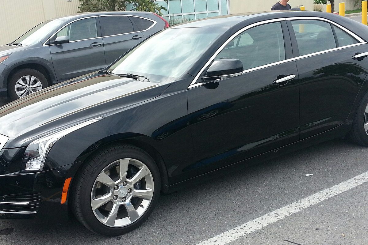 A black Cadillac ATS sedan with silver multi-spoke wheels, parked in a lot next to a gray crossover