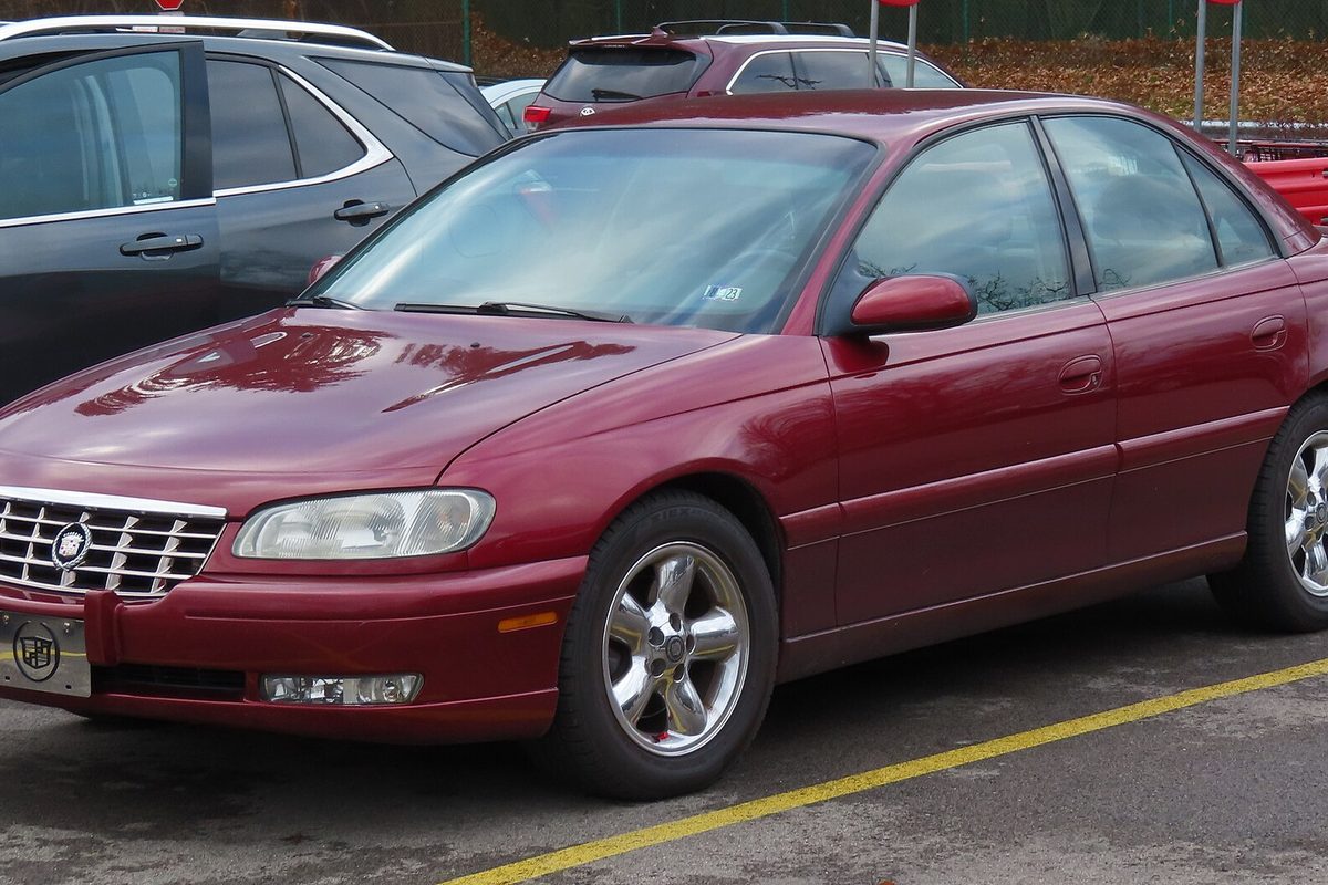 A cranberry red Cadillac Catera sedan with chrome five-spoke wheels, parked in a lot beside an SUV