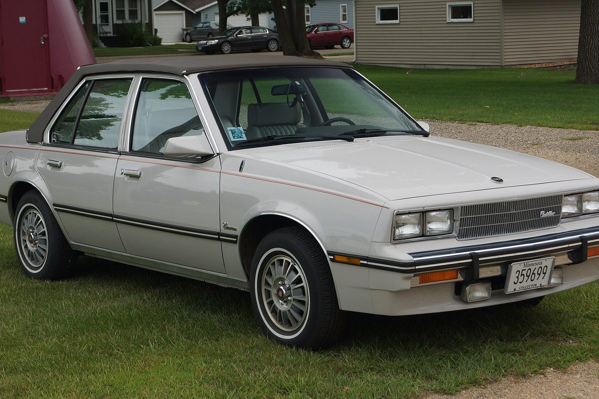A silver Cadillac Cimarron four-door sedan with dark pinstripes and wire-style wheel covers, parked on a lawn