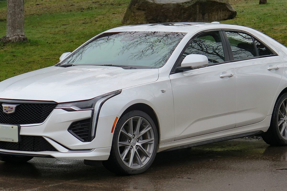 A white Cadillac CT4 sedan, front three-quarter view, on wet pavement beside a grassy park