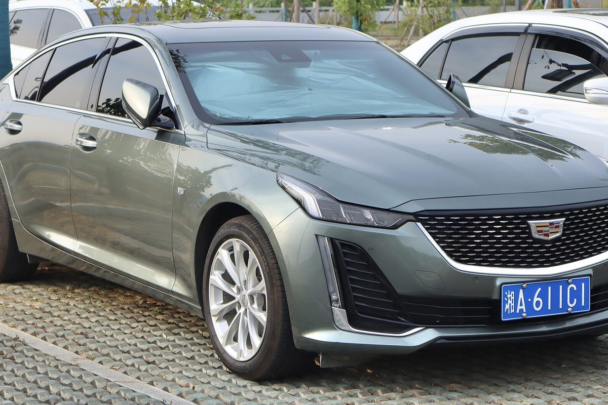 A gray-green Cadillac CT5 sedan with a black mesh grille, parked on a paved lot beside other cars