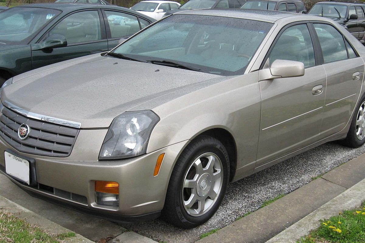 A champagne first-generation Cadillac CTS sedan parked at a curb on gravel, seen from the front and passenger side