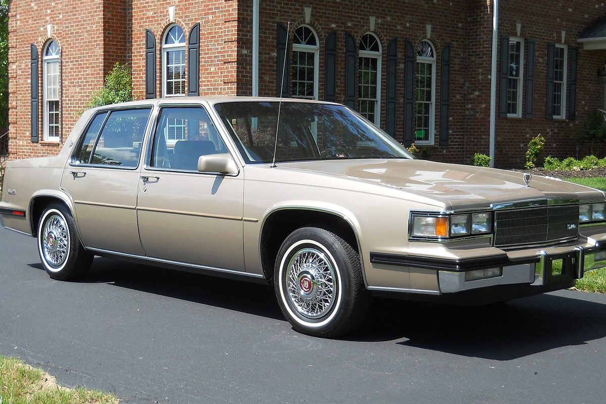 A champagne-gold 1985 Cadillac Sedan DeVille with wire wheels and whitewall tires, parked in front of a brick house