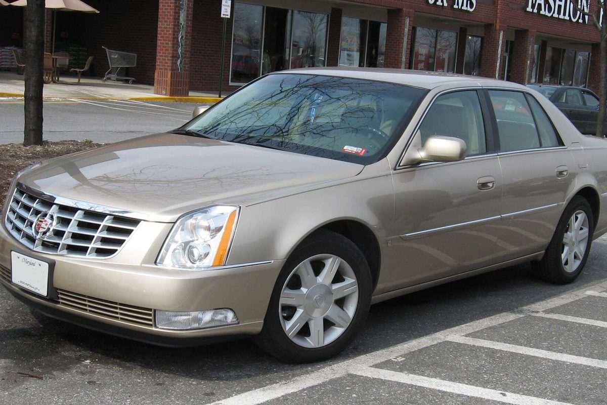 A champagne Cadillac DTS sedan parked in a shopping-center lot, seen from the front and passenger side