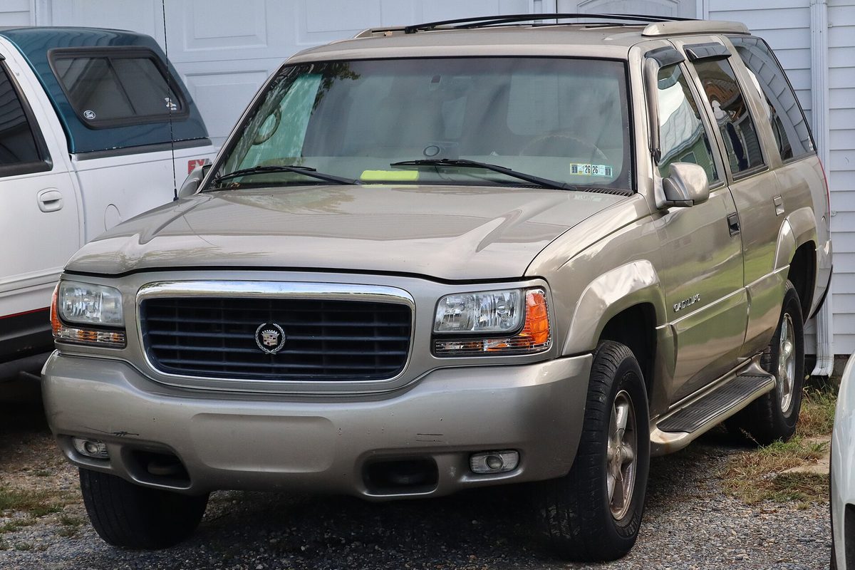 A Silver Sand 1999 Cadillac Escalade, the first-generation SUV, parked on gravel next to a white pickup