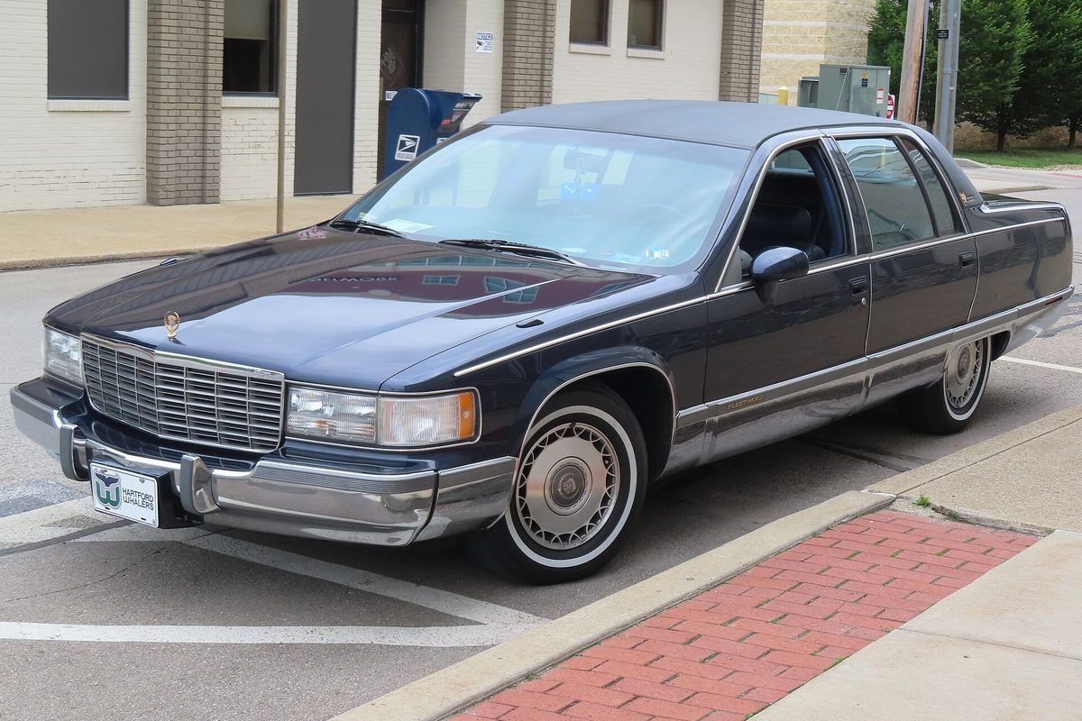 A dark blue Cadillac Fleetwood Brougham sedan with a vinyl roof, whitewall tires and wire wheel covers, parked at a curb