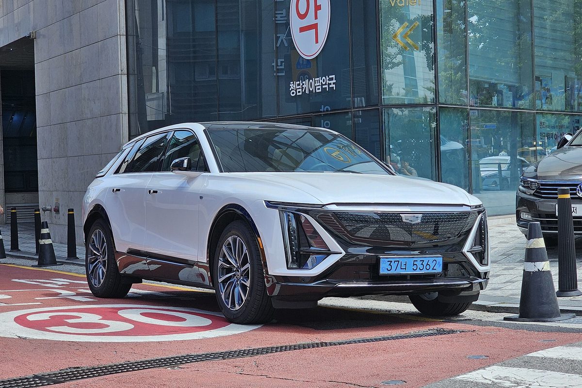 A white Cadillac Lyriq electric SUV with a black lower grille, parked at a city curb in front of a glass office building