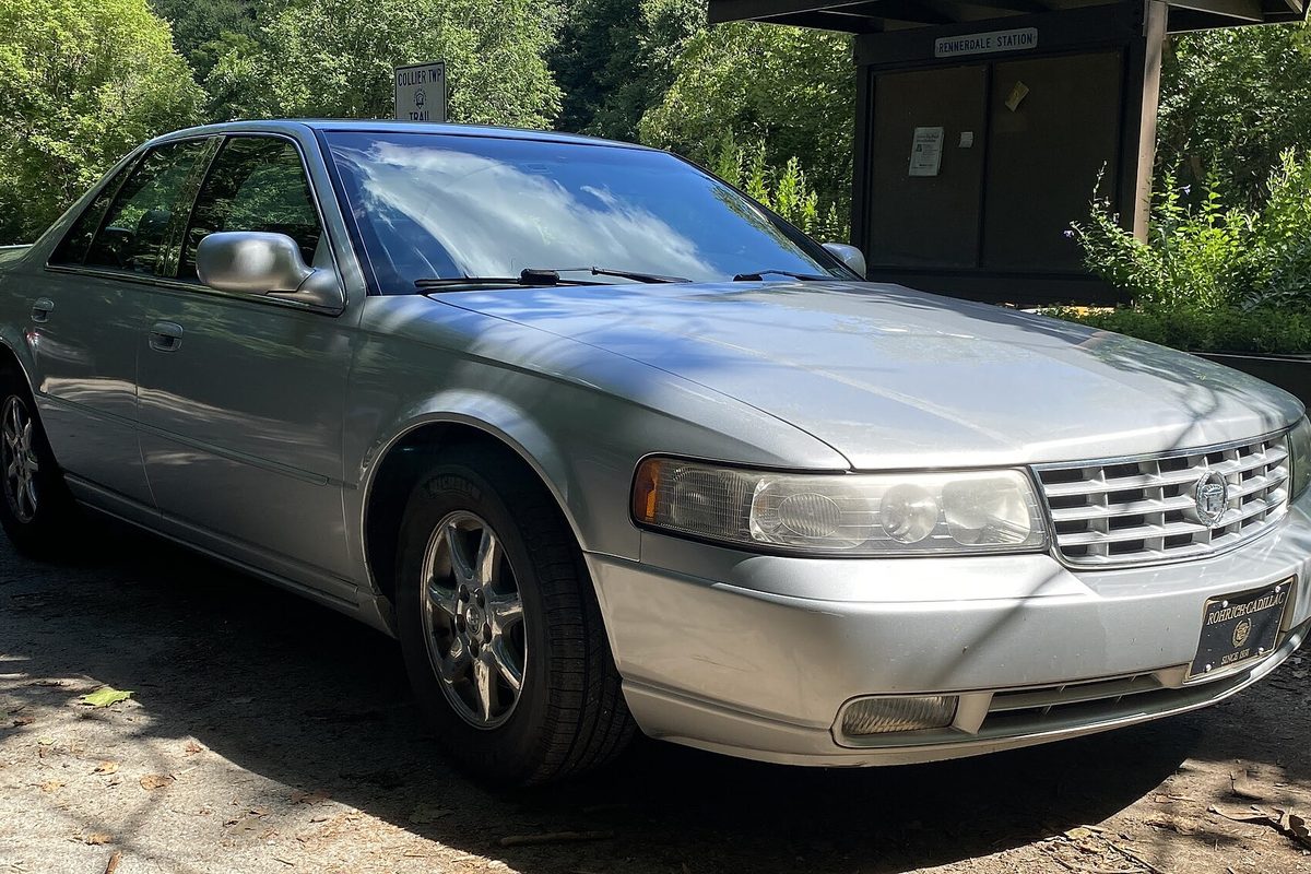 A silver 1999 Cadillac Seville STS sedan parked in dappled shade on a dirt lot, seen from the front