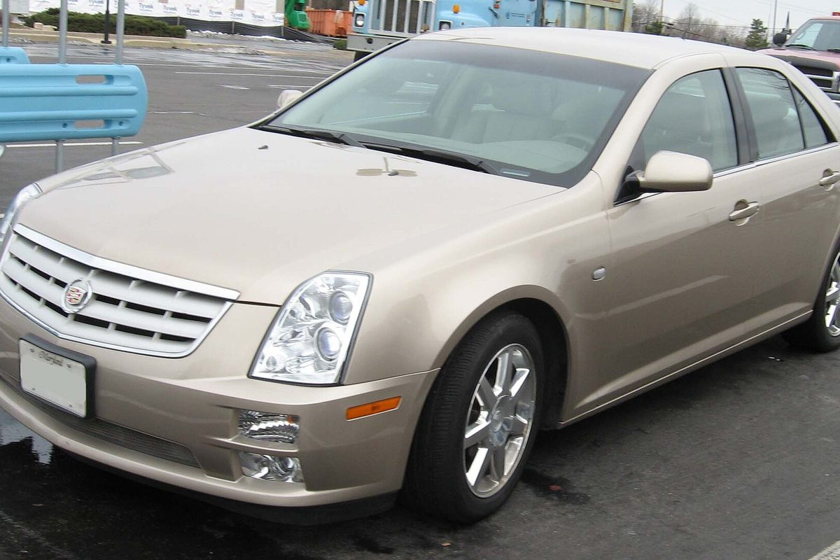 A champagne-gold Cadillac STS sedan with chrome wheels, seen from the front left on a wet parking lot