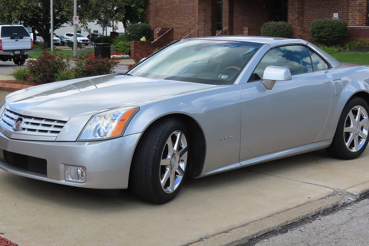 A silver Cadillac XLR two-seater with its roof up, parked on a concrete lot in front of a brick building