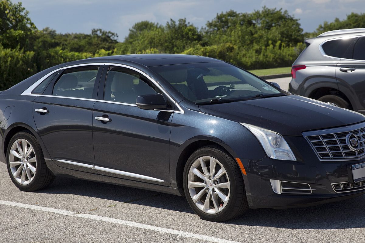 A dark blue-gray Cadillac XTS sedan with silver wheels, seen from the front right in a parking lot by trees