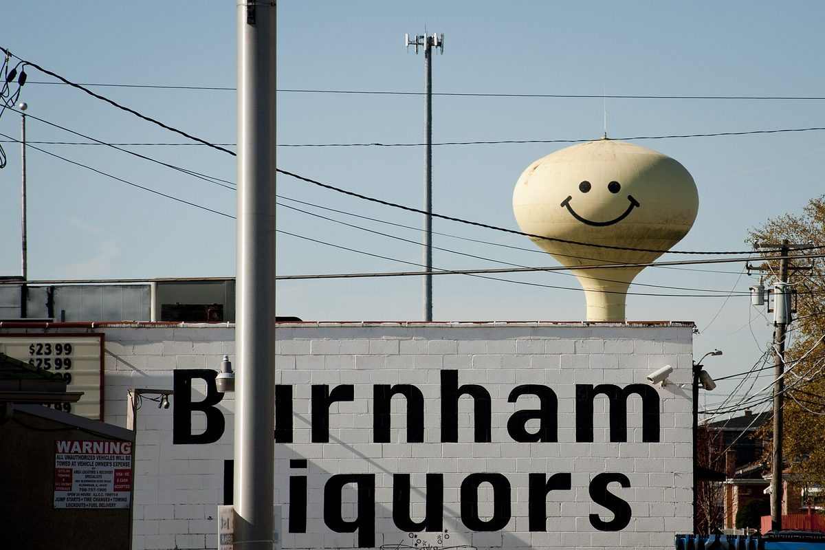 A Calumet City water tower painted with a smiley face, rising behind a white brick building with large black Burnham Liquors lettering