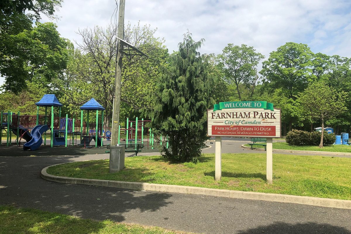 A wooden Welcome to Farnham Park, City of Camden sign on a grassy island beside a playground with blue slides and shade trees