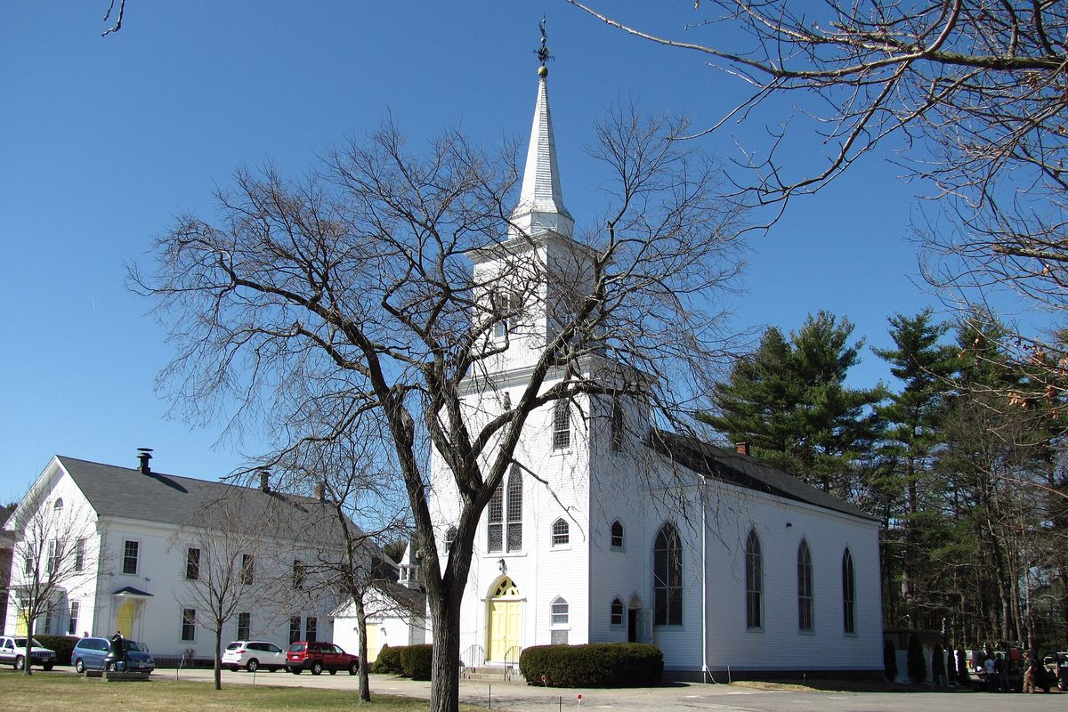 First Parish in Canton Corner, a white church with a tall steeple, seen through bare trees under a blue sky with cars parked beside it