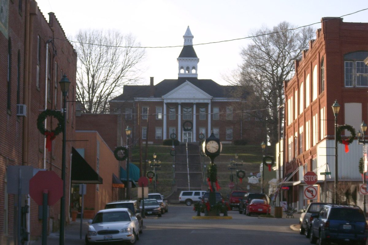 A downtown Cape Girardeau street climbing toward the white-columned City Hall on a hill, with wreaths on the lampposts and cars parked at the curb