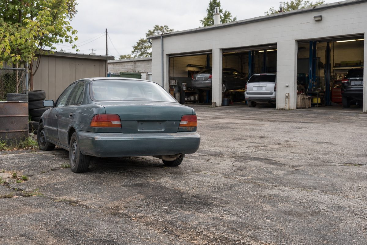 An older green sedan parked on a cracked lot outside a repair garage, with other cars inside its open service bays