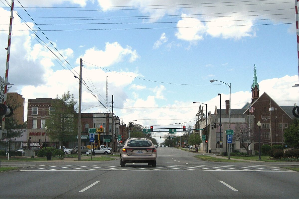 Main Street in Carbondale seen from behind a car at a railroad crossing, with the gates raised, brick buildings and a church steeple ahead