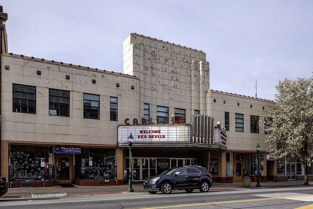 The cream art deco front of the Carlisle Theatre with a marquee reading Welcome Red Devils, a dark SUV at the curb and a flowering tree at the right