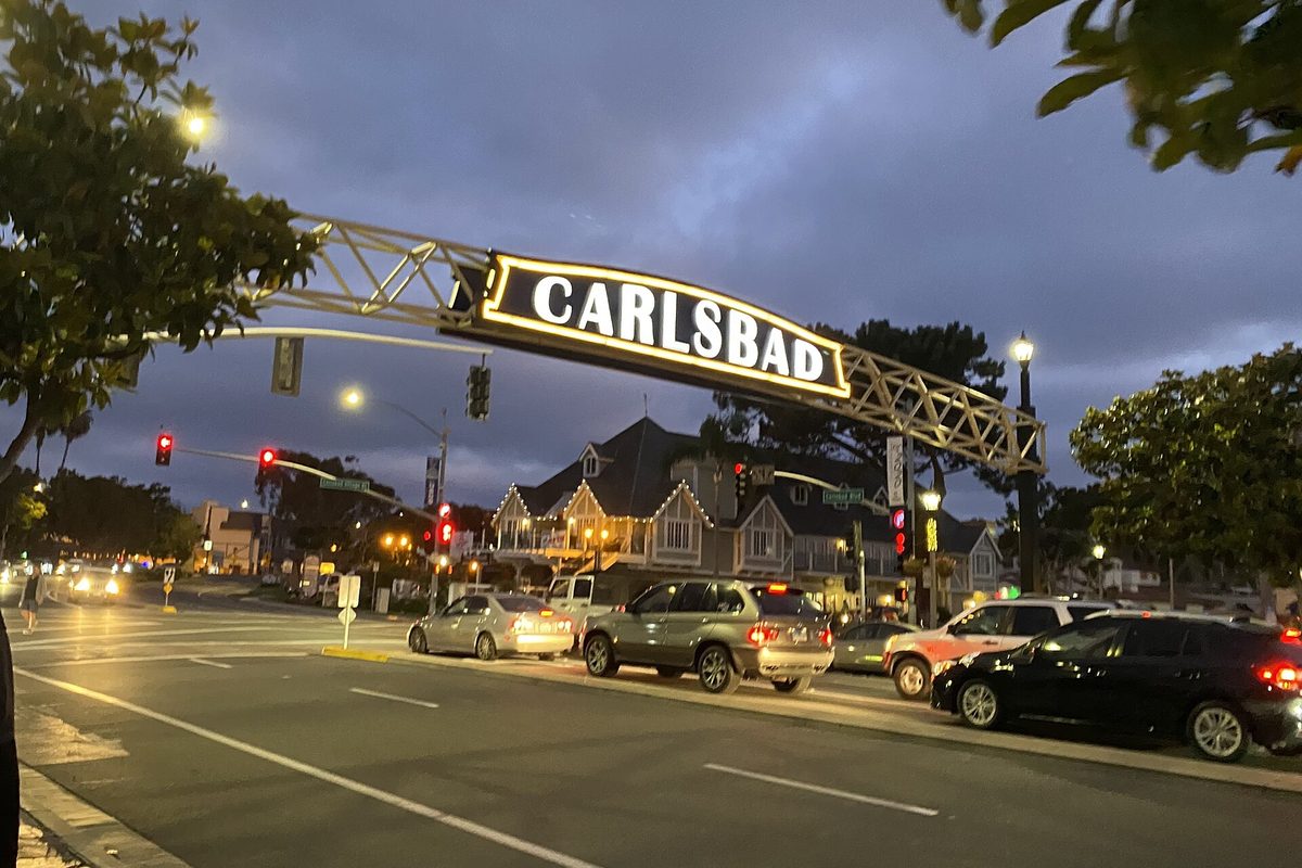 The lit Carlsbad sign arching over Carlsbad Boulevard downtown at dusk, with cars stopped at a red light and a gabled building behind