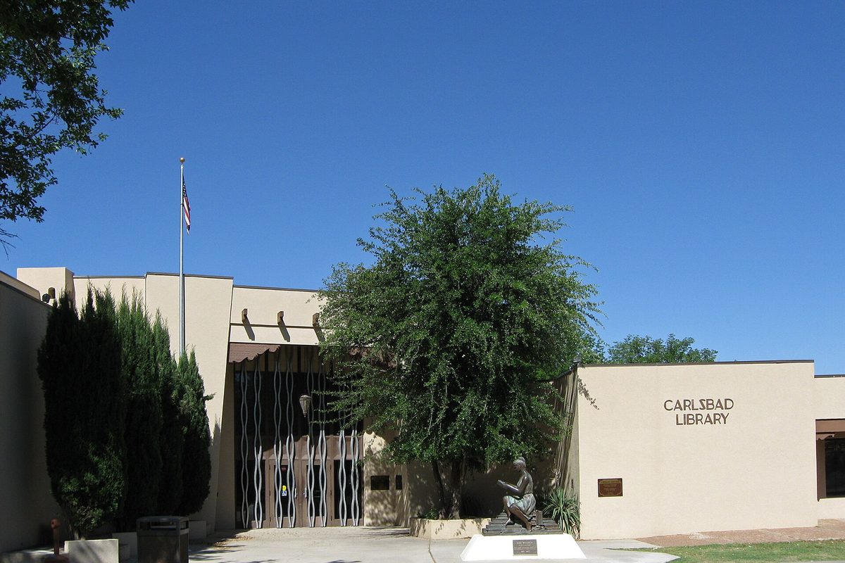 Front of the Carlsbad Public Library, a tan stucco building with a large tree, a flagpole and a bronze statue by the entrance