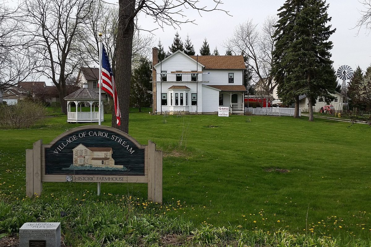 A Village of Carol Stream Historic Farmhouse sign on the lawn in front of the white Stark farmhouse, with a flag, a white gazebo and a windmill