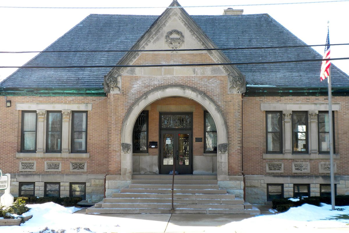 Library Hall in Carpentersville, a brick building with a stone arched entrance, slate roof and an American flag, with snow on the ground