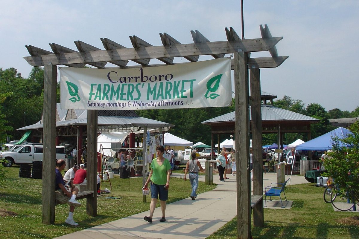 A wooden pergola with a Carrboro Farmers' Market banner over the path, shoppers walking toward white market tents on a sunny day