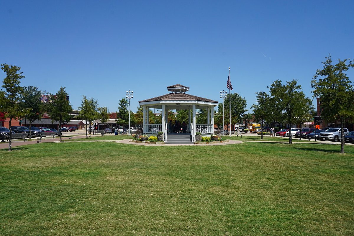 A white gazebo on the lawn of Carrollton Square under a clear blue sky, with an American flag and parked cars along the square