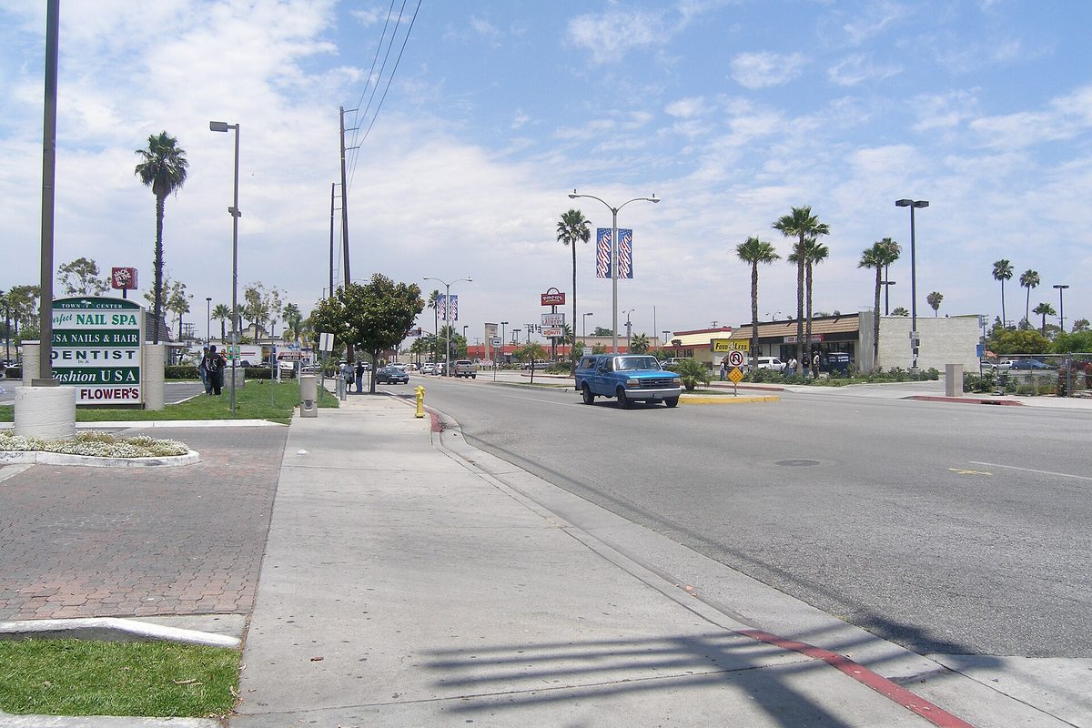 A wide Carson boulevard lined with palm trees and tall streetlights, a blue pickup passing a shopping-center sign under a partly cloudy sky