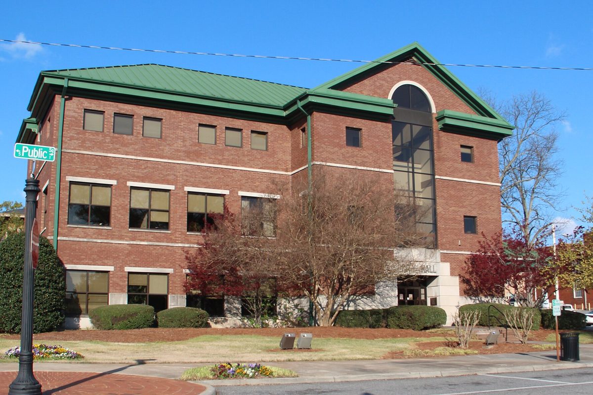 The three-story red-brick Cartersville City Hall with a green metal roof and a tall arched glass entry, a Public Square street sign at left