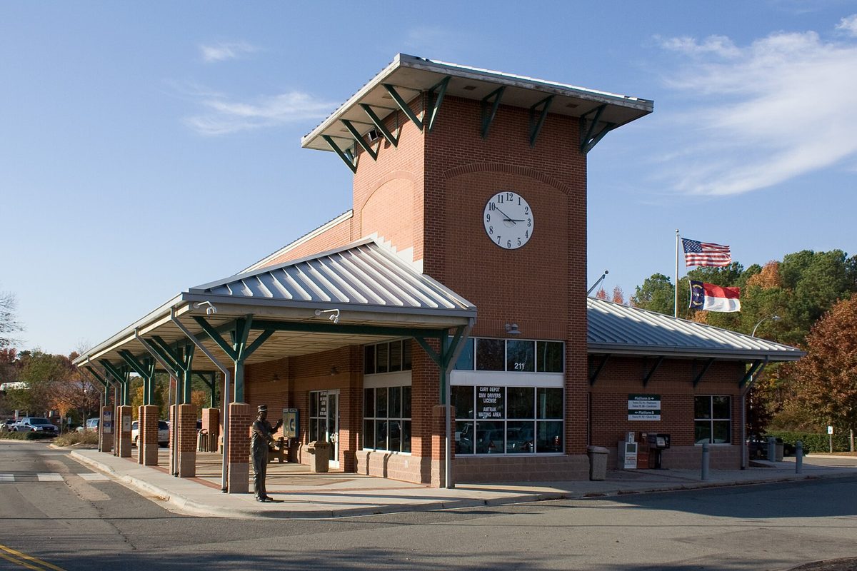 Cary's red-brick train station with a square clock tower, green metal canopy and U.S. and North Carolina flags under a clear blue sky