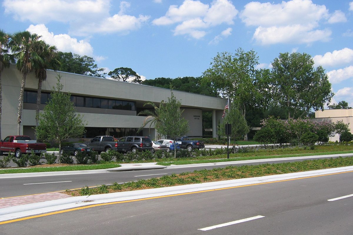 Casselberry City Hall, a low two-story building with dark windows, palm trees and cars in front, beside a landscaped road median