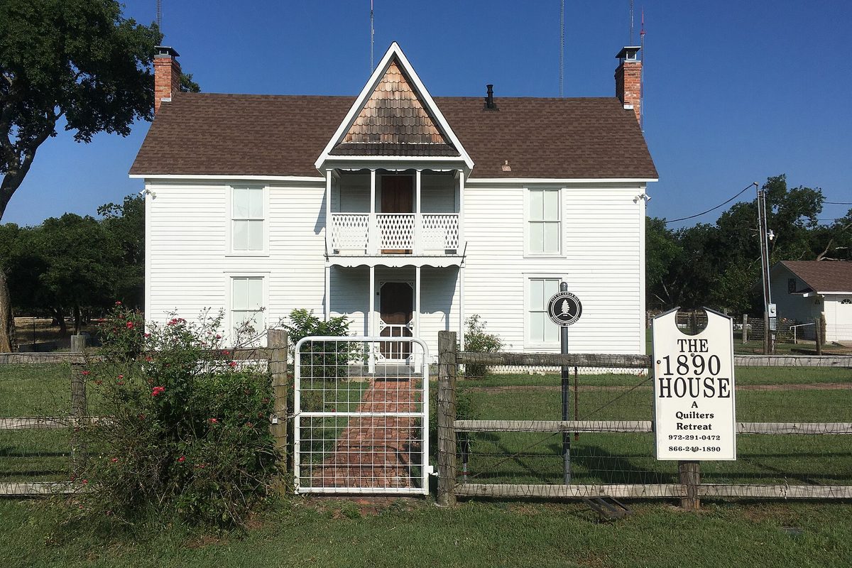A white two-story historic farmhouse in Cedar Hill used as a quilters' retreat, with a brick path, a wire gate in a wooden fence and a sign out front