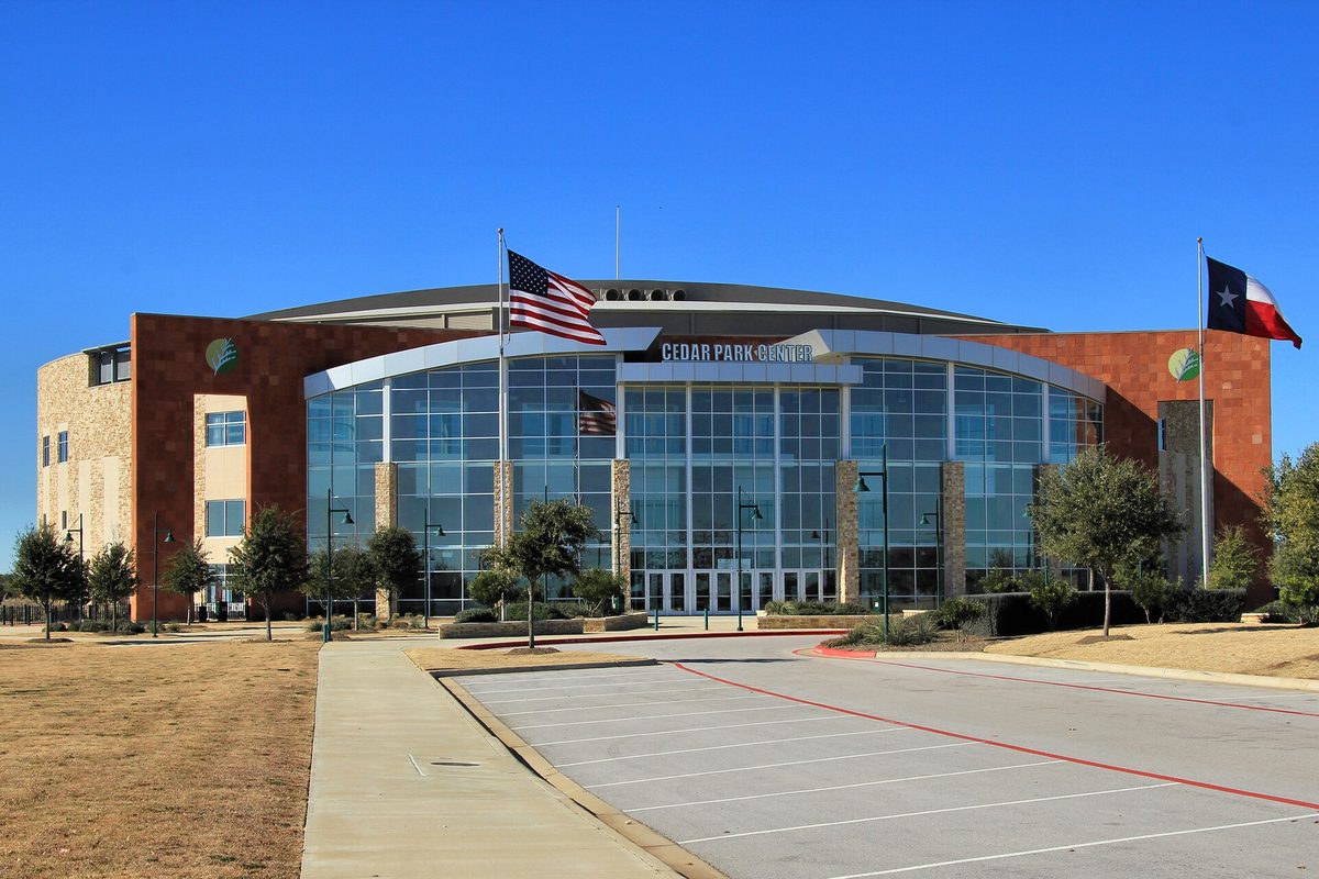 Cedar Park Center, a glass-fronted arena with stone and red brick wings, flying U.S. and Texas flags over an empty parking lot on a clear day