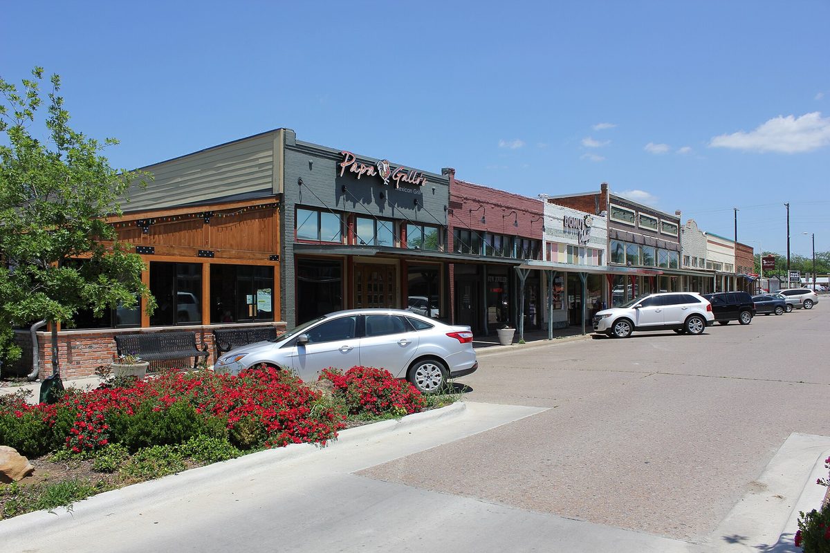 Brick and stone storefronts on a downtown Celina street, with a silver sedan parked beside a bed of red flowers under a clear blue sky