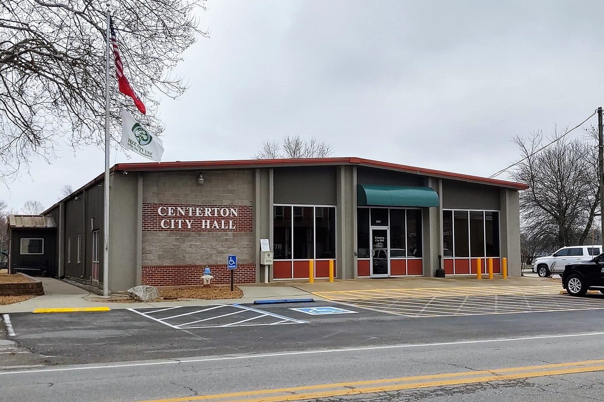 Centerton City Hall, a one-story building with a brick name sign, a flagpole and a green awning over the door, beside an empty parking lot on a gray day