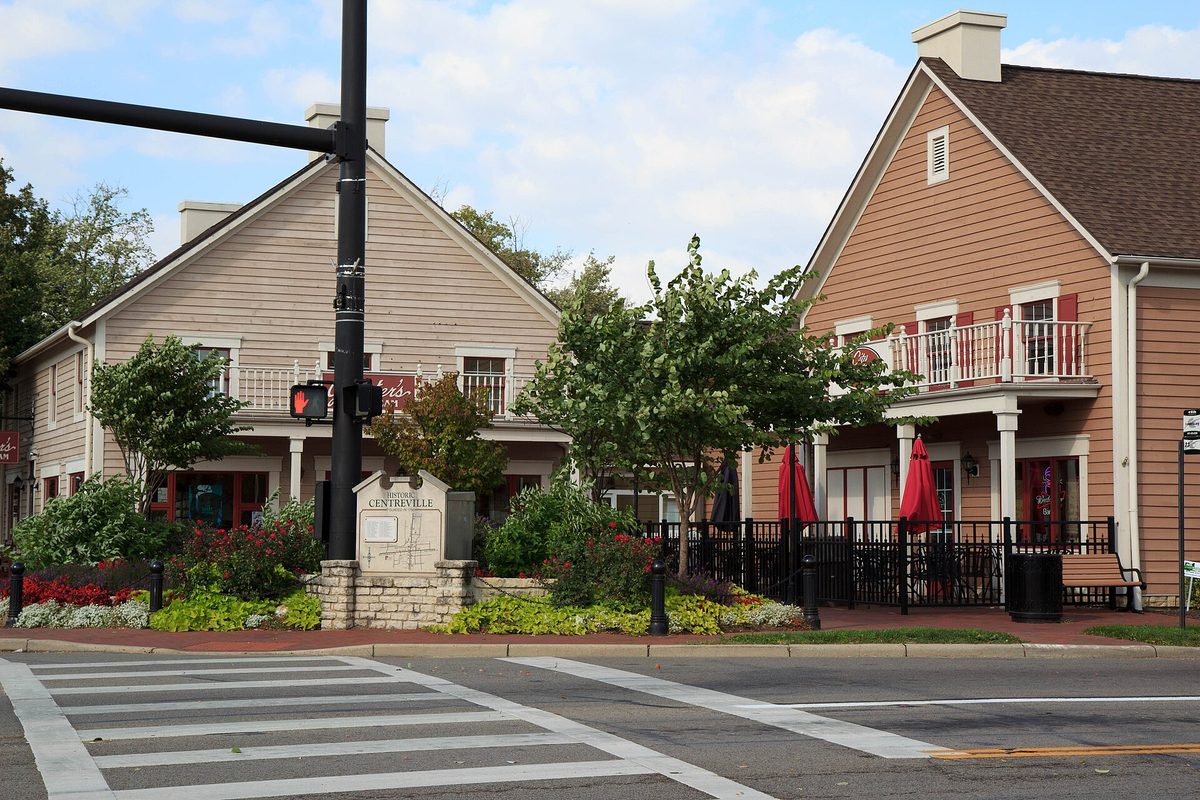 Two restored clapboard buildings with balconies and red umbrellas in the Centerville Historic District, behind a stone Centerville marker, a traffic signal and a crosswalk