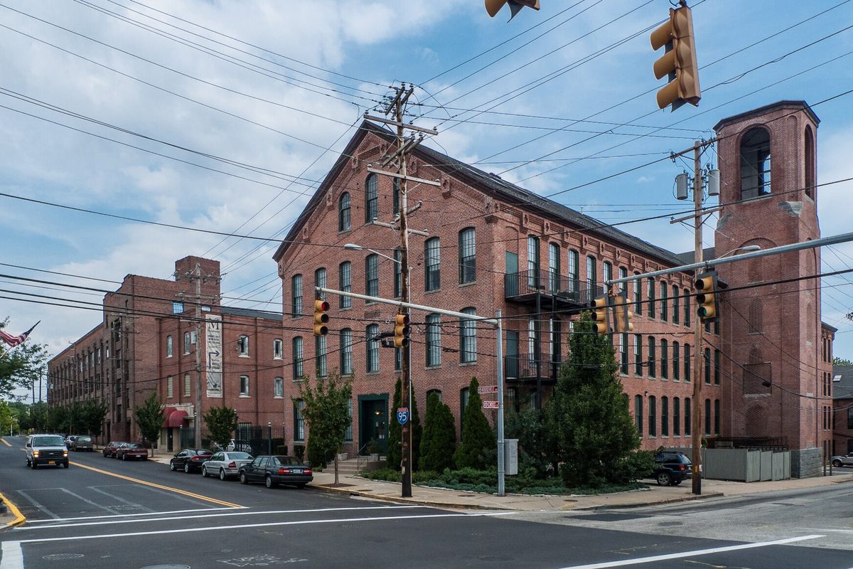 A long red-brick mill building with a square bell tower on a Central Falls street corner, traffic lights overhead and cars parked along the curb