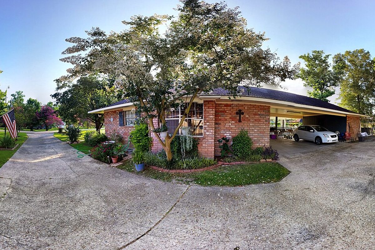 A brick ranch house in Central with a carport and a white car, an American flag and flowering trees in the yard