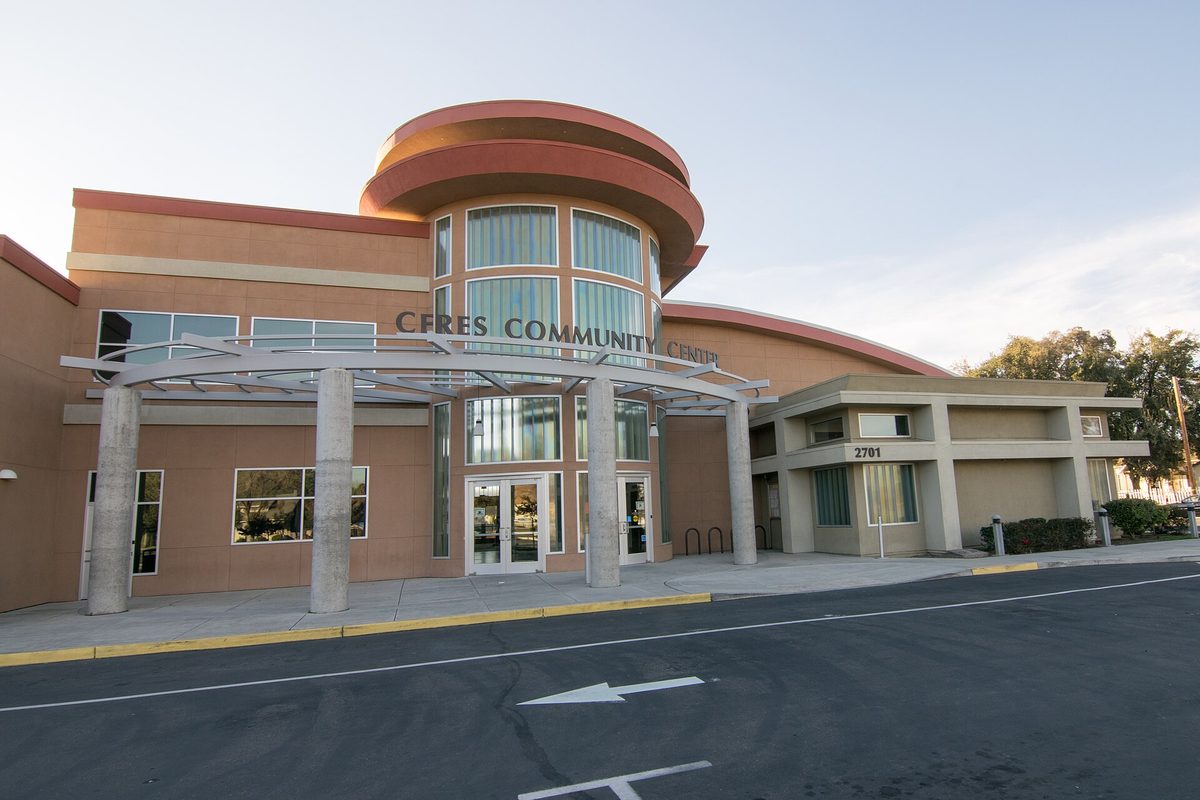 The Ceres Community Center, a tan and terracotta building with a rounded glass entrance tower and a column-lined canopy, seen across its parking lot
