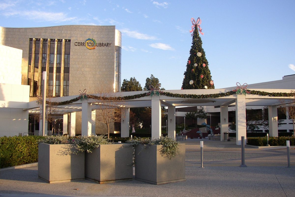 The Cerritos Library, a curved stone and glass building, with a tall decorated Christmas tree behind a white colonnade hung with garland and a row of planters in front