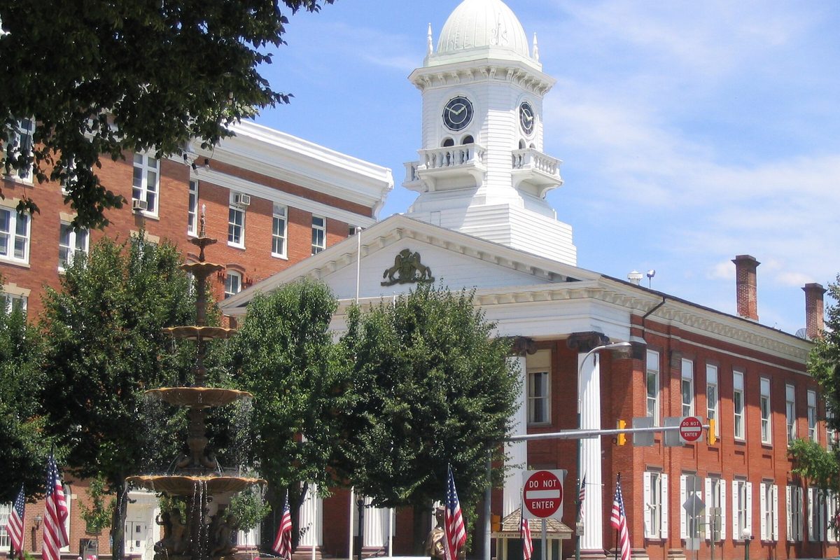 Memorial Square in Chambersburg: a white clock-tower cupola above a columned brick building, a tiered fountain and trees in front, and American flags along the sidewalk
