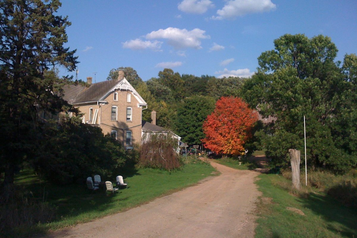 The cream-brick Albertine and Fred Heck House in Chanhassen beside a dirt lane, with a bright red-leafed tree and lawn chairs on the grass