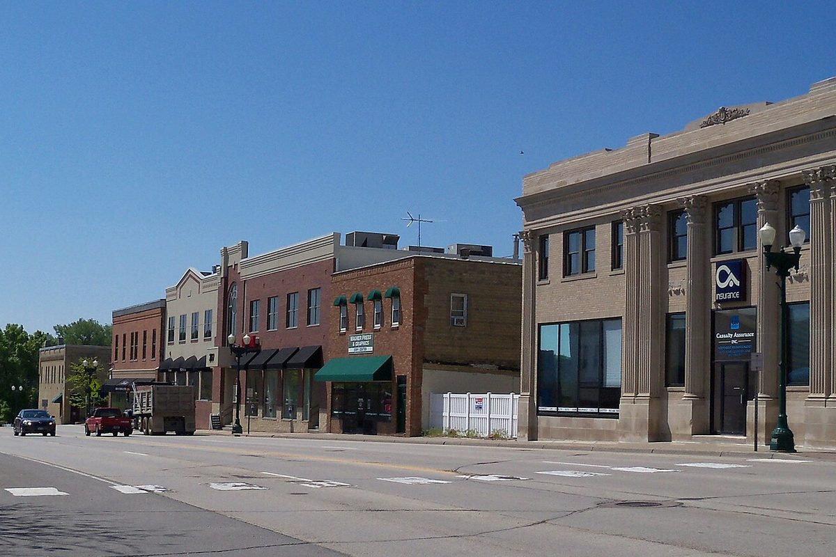 A downtown Chaska street with a row of brick storefronts and green awnings, a pale stone bank building with tall columns on the corner and a few pickups at the curb under a clear blue sky