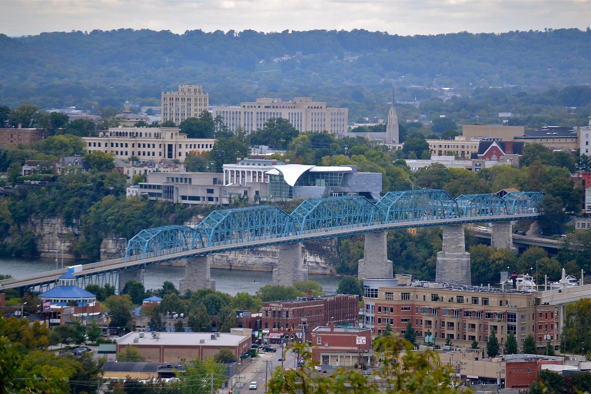 A blue steel truss bridge crossing into downtown Chattanooga, seen from Stringer's Ridge, with wooded ridges behind the office towers