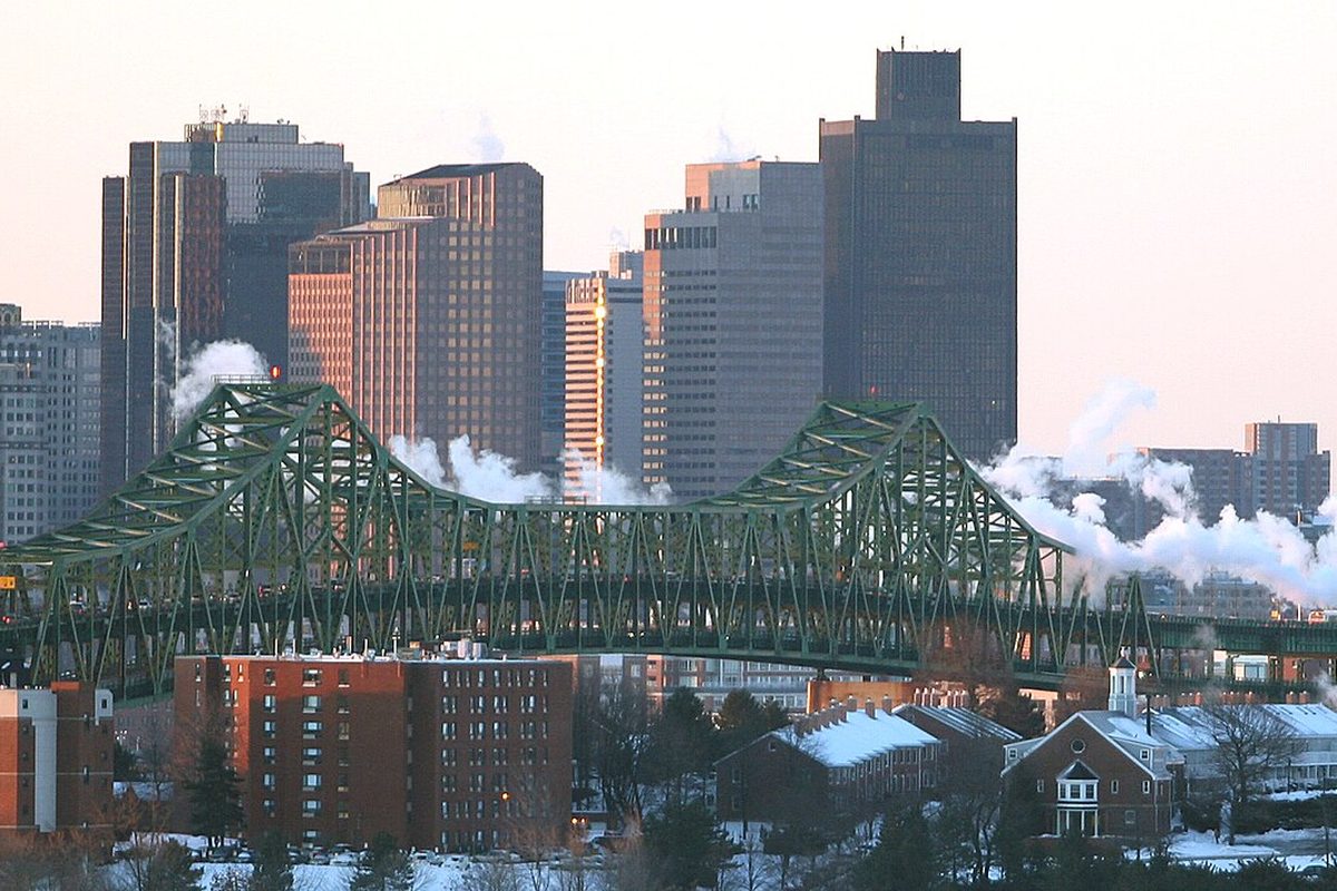 The green steel truss of the Tobin Bridge in front of the Boston skyline on a winter evening, with snow on the roofs of brick buildings below
