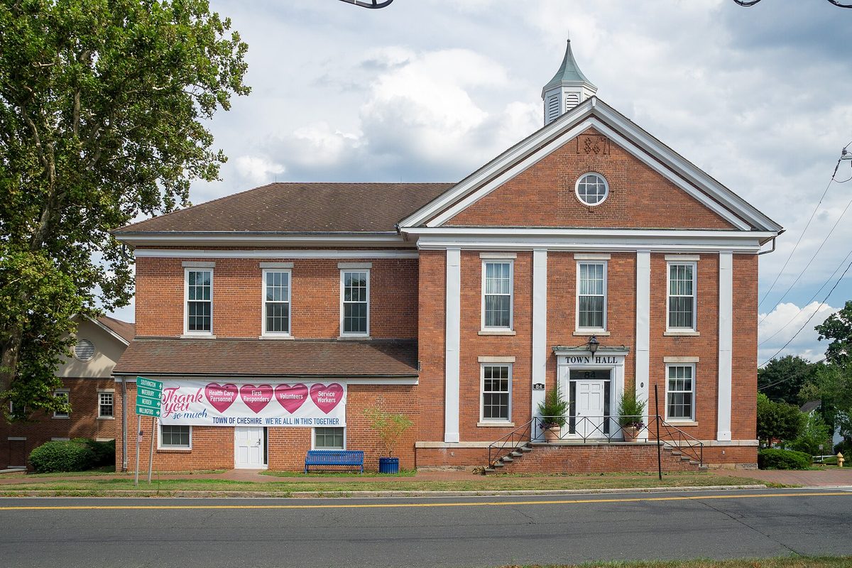 Cheshire Town Hall, a red brick building with white pilasters and a small cupola, with a thank-you banner on its side wing and a blue bench out front