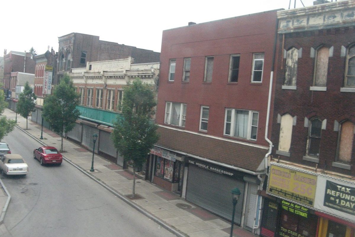 Brick storefronts along Avenue of the States in Chester seen from a train, with young street trees, shuttered shop fronts and two cars parked at the curb